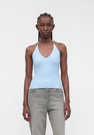 Jeune femme aux cheveux tressés portant un haut dos nu bleu clair et un jean gris, debout devant un fond clair uni.