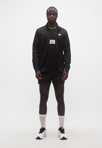 Άνδρας που φοράει μαύρα αθλητικά ρούχα Puma, μαύρη κορδέλα στο κεφάλι, λευκές κάλτσες και λευκά αθλητικά παπούτσια, στέκεται μπροστά σε ένα απλό λευκό φόντο.