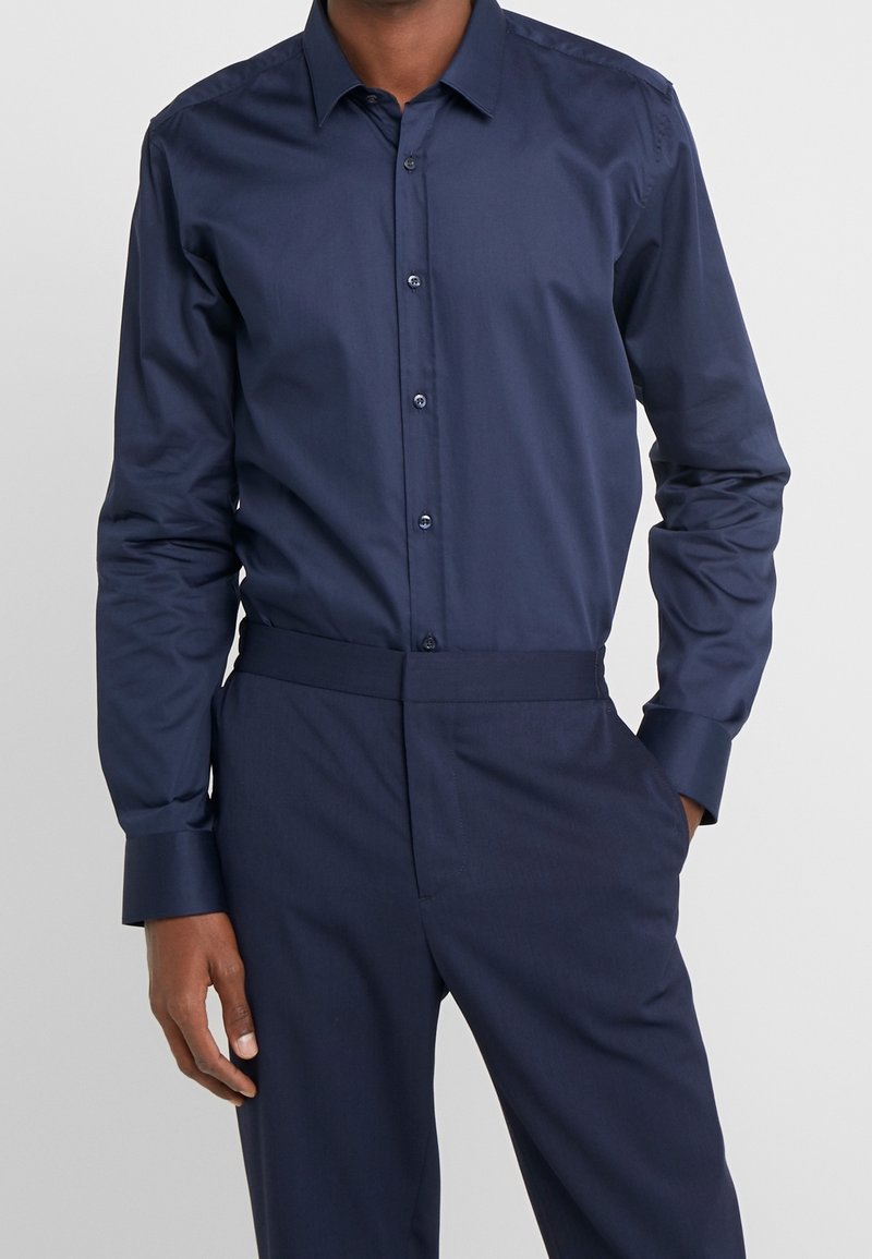 Homme portant une chemise ajustée à manches longues bleu foncé boutonnée, rentrée dans un pantalon assorti bleu foncé.