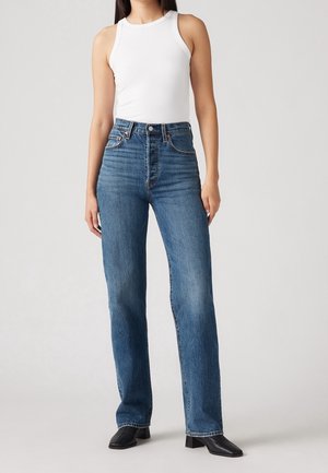 Vrouw met hoge taille blauwe jeans, wit mouwloos geribd topje en zwarte enkellaarsjes met vierkante neus, staand tegen een effen achtergrond.