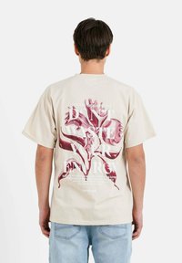 Camiseta de algodón beige con un gran gráfico floral rosa y texto blanco en la parte posterior. Con mangas cortas y un ajuste relajado.