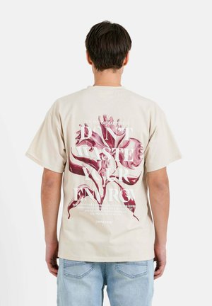 Beige cotton t-shirt featuring a large pink floral graphic and white text on the back. Short sleeves and a relaxed fit.