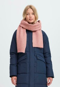 Blondide juustega naine, kellel on seljas tumesinine puffer-jakk ja paks roosa kiudne sall, mis on ümber kaela mähitud, seisab lihtsa tausta ees.