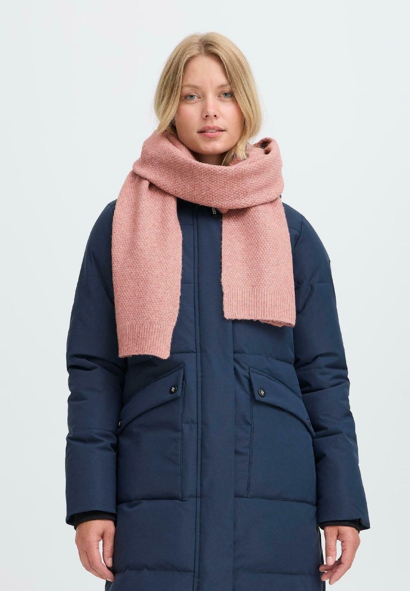 Blondide juustega naine, kellel on seljas tumesinine puffer-jakk ja paks roosa kiudne sall, mis on ümber kaela mähitud, seisab lihtsa tausta ees.