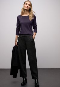 Fioletowa bluzka z długim rękawem w zestawieniu z czarnymi, szerokimi spodniami o wysokim stanie i fakturze. Modelka trzyma czarną kurtkę, ma na sobie czarne botki.