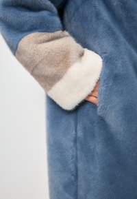 Palton din blană sintetică de culoare albastră deschisă, cu accente bej și alb la manșetă. Finisaj texturat cu detalii la buzunare laterale, design neted și pufoș.
