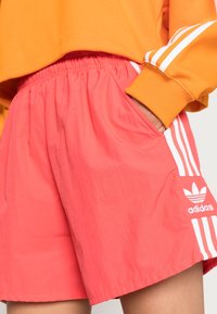 Person, der korallenfarbene Adidas-Shorts mit weißen Streifen und Logo trägt, die Hand in der Tasche, kombiniert mit einem sichtbaren ärmel eines orangefarbenen Sweatshirts.