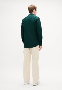 Donkergroen overhemd met knopen, gezien vanaf de achterkant, gecombineerd met crèmekleurige losse broek. Eenvoudige, gladde stof met een standaard kraag.