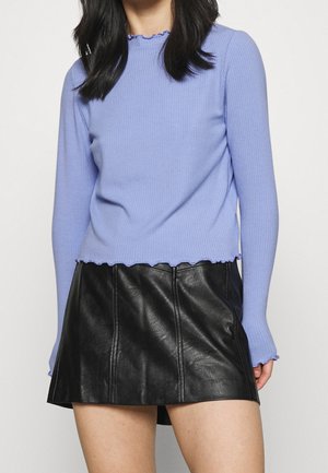 Nainen, joka pukeutunut vaaleanpunaiseen, ribbattuun, pitkähihnaiseen paitaan, jossa on lehtireunukset, ja mustaan nahkaiseen minihameeseen, kuvattuna kaulasta reiteen.