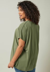 Top verde oliva a maniche corte realizzato in tessuto morbido, con un design ampio e cucitura centrale sul retro, abbinato a jeans blu.