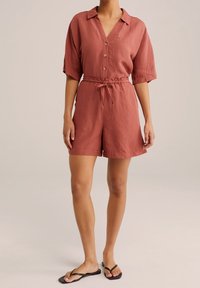 Terakotový lněný overal s límečkem, zapínáním na knoflíky vpředu, krátkými rukávy, elastickým pasem a šňůrkou. Má volný střih a kraťasy.