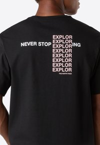 Man som bär en svart t-shirt med vit och rosa text "Never stop exploring" ordnad vertikalt och horisontellt på ryggen.