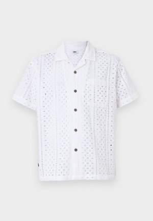 Camisa branca de manga curta com padrões florais recortados, frente abotoada, bolso no peito e botões castanhos. Feita de um tecido leve.