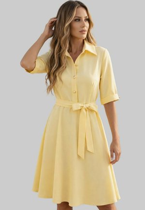 Femme aux longs cheveux ondulés portant une robe jaune boutonnée avec des manches trois-quarts et une ceinture nouée à la taille, regardant sur le côté sur un fond uni.