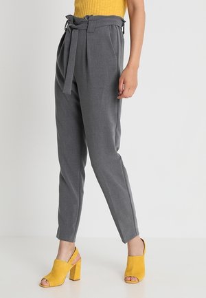 Vrouw draagt grijze, hooggetailleerde taps toelopende broek met stoffen riem, gecombineerd met gele open-toe blokhak schoenen en een geel geribd topje.