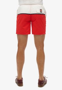 Shorts de bain rouges avec un panneau blanc et des bordures noires, dotés d'une poche arrière avec une étiquette logo. Fabriqués en matériau léger et à séchage rapide.