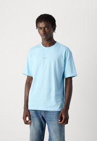 Άνδρας με ανοιχτό μπλε μπλούζα BOSS και μπλε τζιν στέκεται μπροστά σε απλό ανοιχτό γκρι φόντο, κοιτάζει μπροστά με ο�υδέτερη έκφραση.