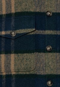 Niebiesko-zielona wełniana tkanina w kratę z kieszenią na piersi, ozdobiona ciemnymi guzikami i miękką, teksturowaną powierzchnią, podkreśloną poziomymi paskami.