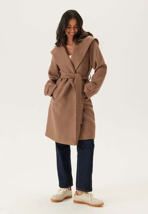 Femme souriante, portant un manteau marron ceinturé, un jean bleu foncé et des baskets blanches, se tenant devant un fond clair.
