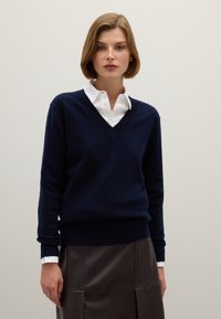 Marineblauwe wollen V-hals trui met lange mouwen, gecombineerd met een wit overhemd met kraag, geplaatst tegen een neutrale achtergrond. Glad oppervlak, casual ontwerp.