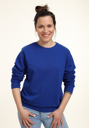 Sweatshirt bleu avec un col rond, des manches longues et des poignets côtelés ; associé à un jeans bleu clair. Texture lisse, coupe décontractée.