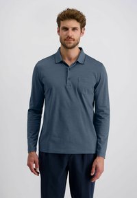 Mann in einem langärmeligen blauen Polohemd und dunklen Hosen, der vor einem einfachen hellgrauen Hintergrund steht.