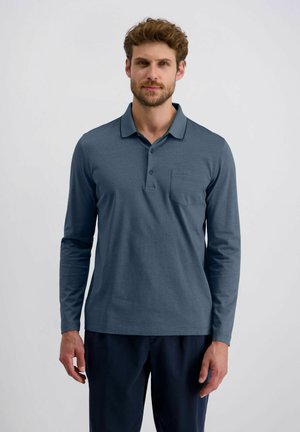 Man in een donkerblauwe poloshirt met lange mouwen en een donker pant stond tegen een effen lichtgrijze achtergrond.