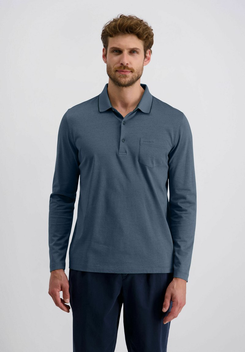 Mann in einem langärmeligen blauen Polohemd und dunklen Hosen, der vor einem einfachen hellgrauen Hintergrund steht.