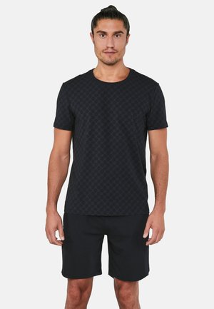 Mand med mørkt hår i en knold iført en sort mønstret kortærmet skjorte og sorte shorts, stående mod en ensfarvet hvid baggrund.