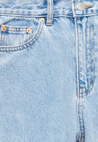 Gros plan sur un jean en denim bleu clair montrant la poche avant, la passants de ceinture, le bouton et les détails visibles de couture orange.