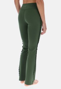 Zielone legginsy z szerokimi nogawkami, o gładkiej teksturze, z falbaniastymi detalami po bokach i wysokim stanem. Materiał wydaje się elastyczny i lekki.