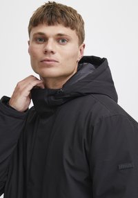 Chaqueta negra con capucha, collar alto y textura suave. Cuenta con una etiqueta de la marca discreta en la manga y un ajuste ceñido alrededor del cuello.