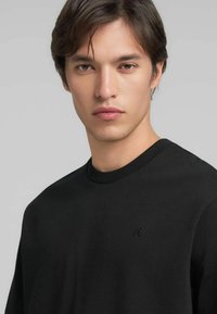 Felpa nera con collo a girocostina, realizzata in un tessuto morbido. Presenta un logo in rilievo sottile sul petto. Tessuto liscio e vestibilità comoda.