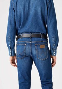 Blaue Jeans aus Denim mit einem braunen Lederpatch, kontrastierendem Stitching und einem schwarzen Gürtel. Langärmliges Hemd aus blauem Denim mit geknöpften Manschetten.