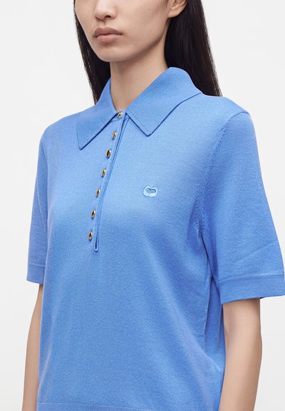 Jeune femme portant un polo bleu clair à manches courtes avec des boutons dorés et un petit logo brodé sur la poitrine.