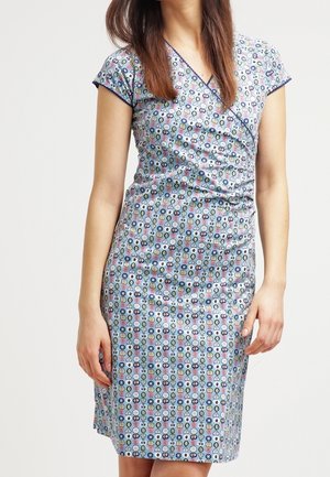 Frau trägt ein eng anliegendes hellblaues Kleid mit kurzen Ärmeln und einem bunten, sich wiederholenden geometrischen Muster, steht mit entspannten Armen an ihren Seiten.