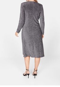 Vestido gris de manga larga con un torso ajustado y una falda acampanada hasta la rodilla. Tela suave y texturizada con un ligero brillo. Llevado con tacones negros con tiras.
