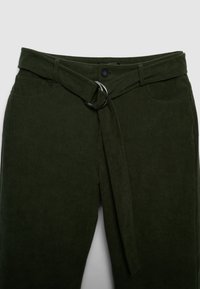 Pantaloni in velluto a coste verde oliva con una larga cintura in tessuto, dotata di fibbia a anello argentata, e due tasche frontali. Superficie testurizzata.