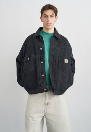 Giovane uomo che indossa una giacca nera larghezza oversize con motivo sopra una maglia verde e pantaloni beige chiaro, in piedi davanti a uno sfondo grigio semplice.