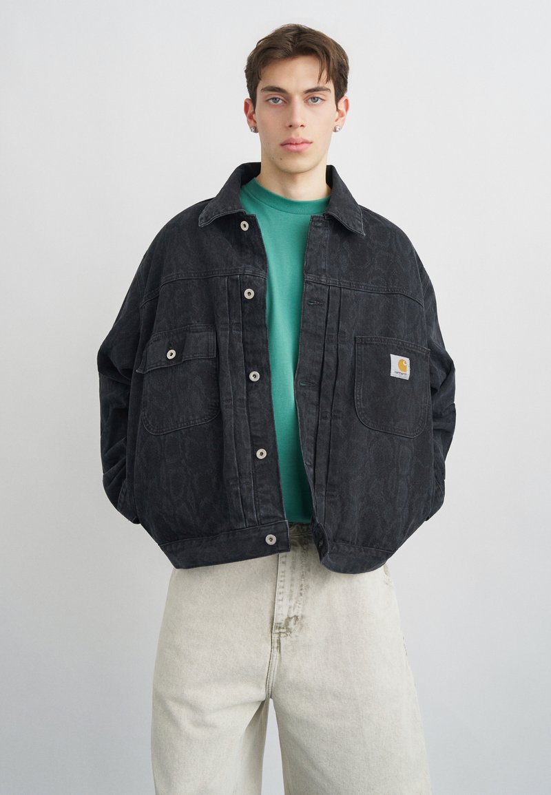 Jonge man die een oversized zwarte jas met patroon draagt over een groen shirt en lichtbeige broek, staand tegen een effen grijze achtergrond.