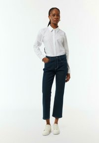 Weißes Button-Down-Hemd mit Kragen, kombiniert mit dunkelblauen hochgeschnittenen Jeans und weißen Turnschuhen. Einfaches Design, klare Linien und figurbetont.
