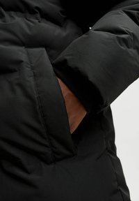 Schwarze Daunenjacke mit brauner Innenauskleidung und einer Seitentasche. Das Material hat eine glatte, steppartige Textur.