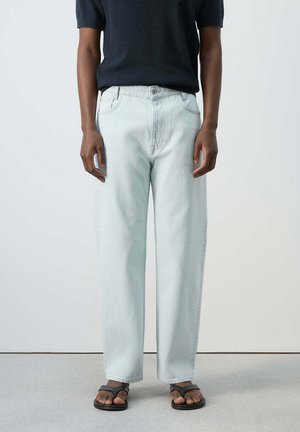 Persona con vaqueros anchos de pierna en azul claro, camiseta negra de punto de manga corta y sandalias negras con correas en los dedos, de pie junto a una pared lisa.