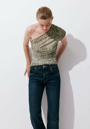 Femme aux cheveux attachés portant un top zébré à une épaule et un jean bleu foncé, debout devant un fond blanc, regardant vers le bas.