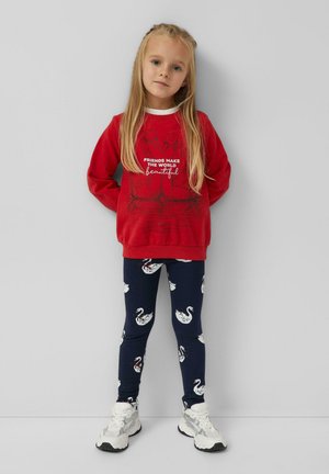 Junges Mädchen steht mit den Händen hinter dem Rücken, trägt einen roten Pullover mit Schwänen und Text, marineblaue Leggings mit weißen Schwan-Mustern und weiße Turnschuhe.