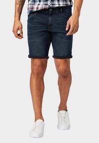 Hombre con pantalones cortos de mezclilla oscuros y enrollados, zapatillas blancas y una camisa de cuadros metida por dentro, de pie con las manos relajadas a los lados.