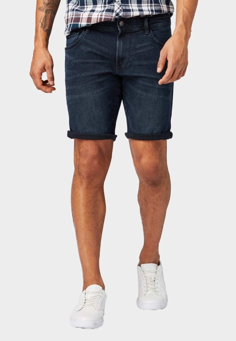 Hombre con pantalones cortos de mezclilla oscuros y enrollados, zapatillas blancas y una camisa de cuadros metida por dentro, de pie con las manos relajadas a los lados.