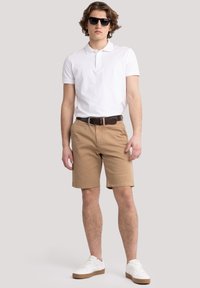 Beige Baumwollkurzshorts, weißes Polohemd und weiße Sneaker. Die Accessoires umfassen dunkle Sonnenbrillen und einen braunen Gürtel. Lässiger Schnitt und Stil.