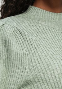 Detailný záber na zelený rebrovaný pletený sveter s diagonálnym ramenným detailom na osobe s tmavými kučeravými vlasmi a stredne tmavou pleťou.