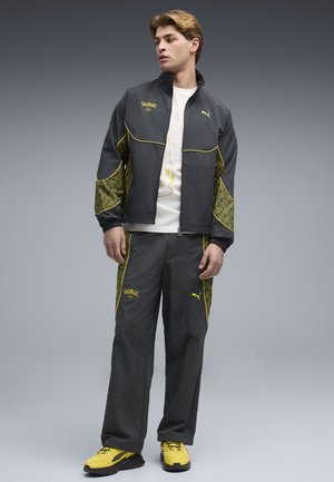 Junger Mann trägt schwarzen und gelben Puma x Pokémon Trainingsanzug mit Reißverschlussjacke, passenden Hosen, weißem Pikachu-T-Shirt und gelben Turnschuhen.